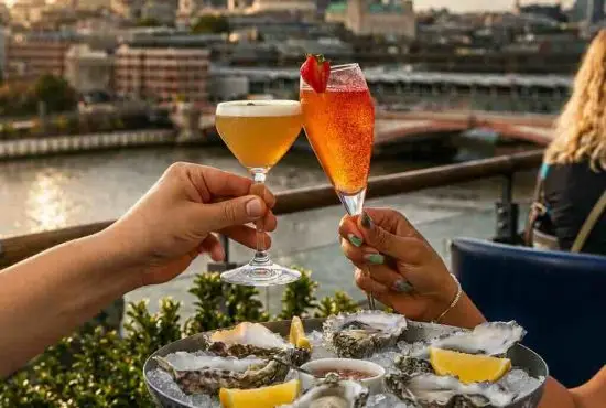 Two hands toasting cocktails above a plate of oysters